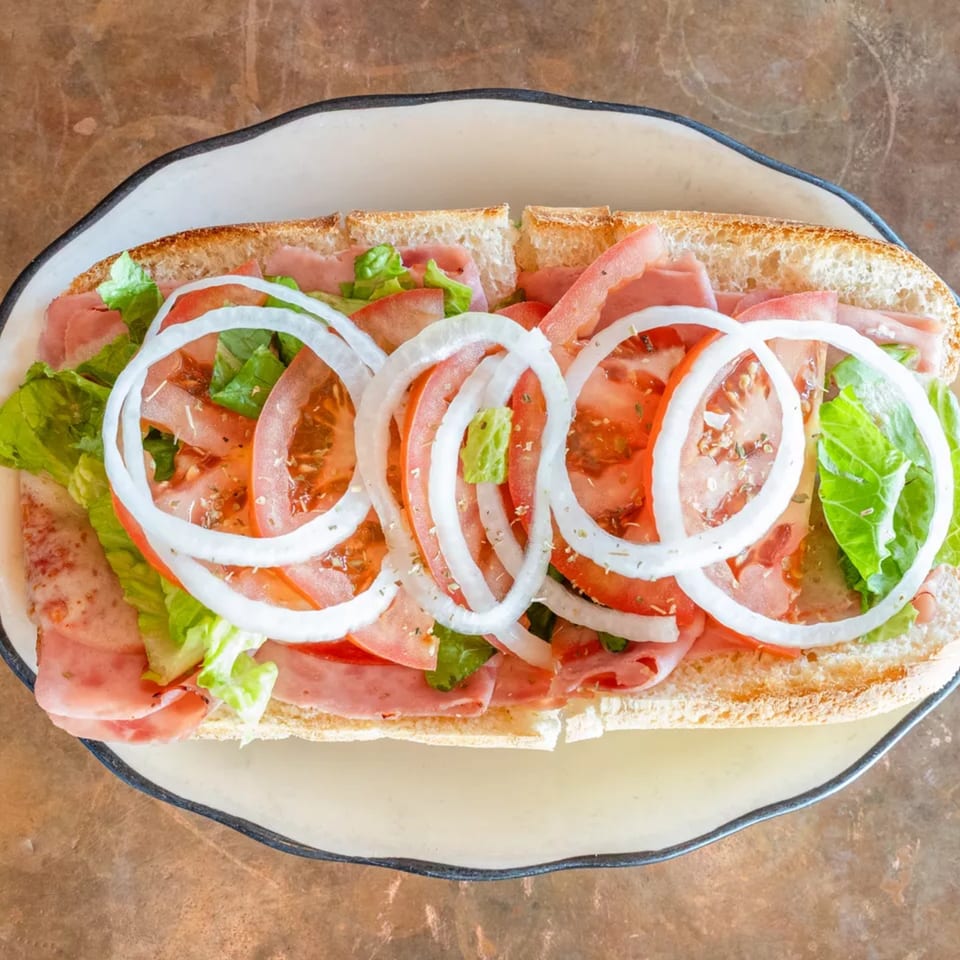 Overhead view of a submarine sandwich with ham, lettuce, tomatoes, and onion rings on a white plate.