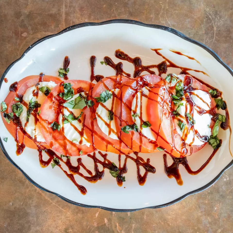 Caprese salad with sliced tomatoes, mozzarella, basil, and balsamic glaze on a white plate.