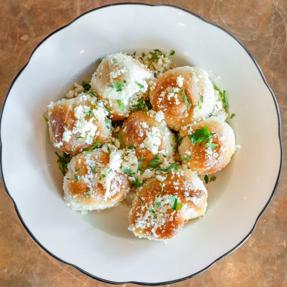 A white bowl filled with golden-brown garlic knots, topped with crumbled cheese and fresh parsley.